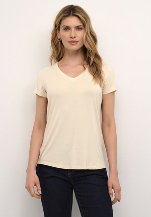 Femme aux cheveux blonds ondulés portant des boucles d’oreilles créoles dorées, un t-shirt crème à manches courtes avec col en V, et un jean bleu foncé, debout devant un fond uni.