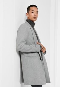 Hellgrauer Wollmantel mit Revers, einem Knopfverschluss und zwei Fronttaschen; getragen über einem dunkelgrauen Rollkragenpullover. Glatte Textur.