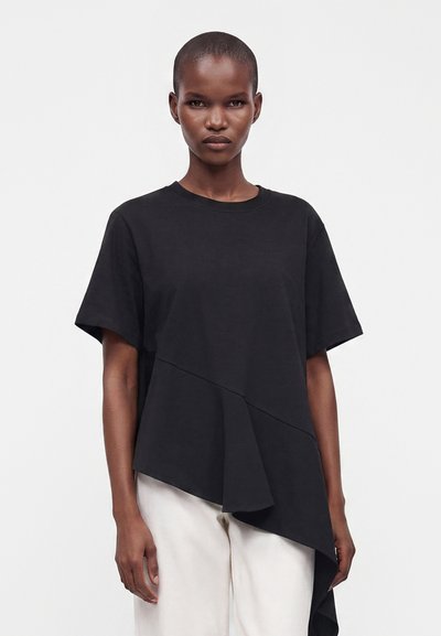 Femme aux cheveux courts portant un haut noir asymétrique à manches courtes et un pantalon blanc, debout sur un fond blanc uni.