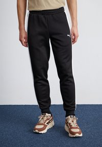 Pantalones de chándal negros con rayas laterales blancas, cinturilla elástica y bolsillos con cremallera; combinados con zapatillas deportivas marrón y crema.