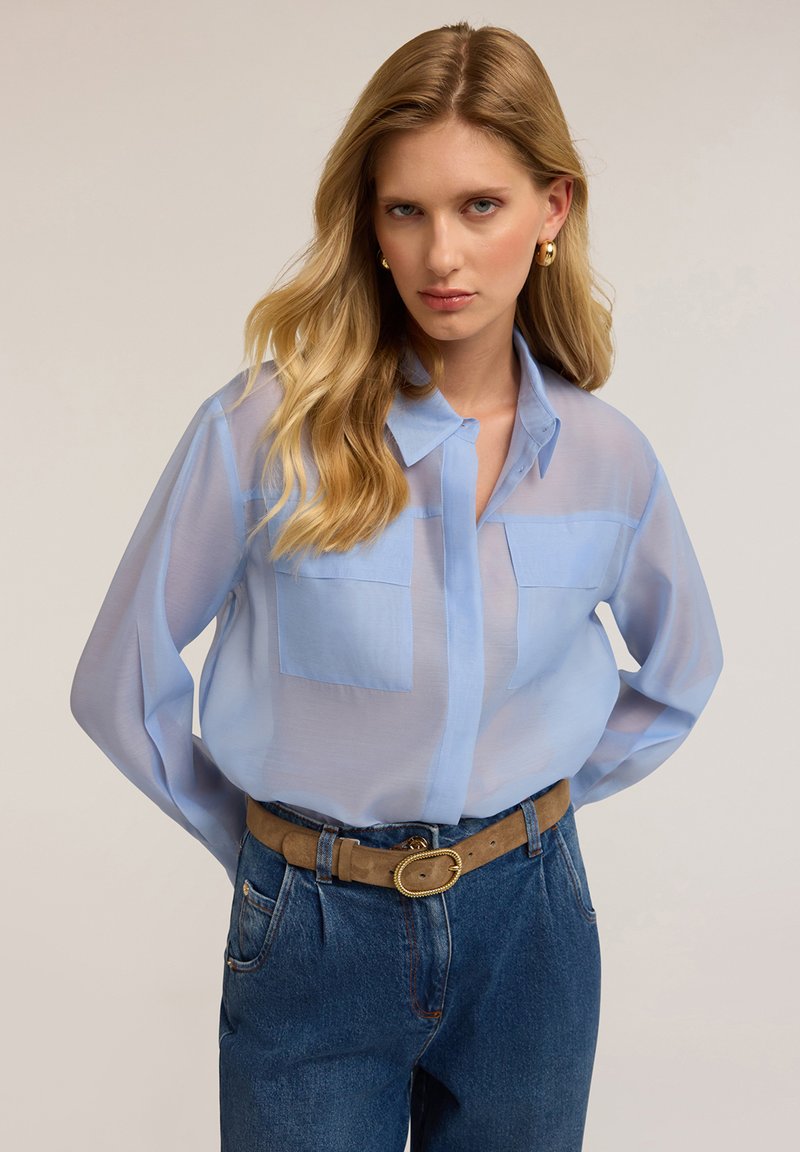 Camicia leggera azzurra trasparente con bottoni e due tasche sul petto, abbinata a jeans scuri a vita alta e una cintura beige con fibbia rotonda.