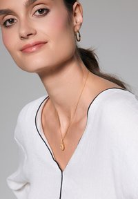 Złoty naszyjnik z zawieszką o ozdobnych detalach na delikatnym łańcuszku, noszony na białej koszuli z czarnym wykończeniem. Małe kolczyki w kształcie obręczy uzupełniają wygląd.