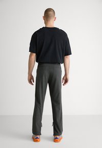 T-shirt en coton noir, pantalon gris ajusté avec fermetures éclair sur les côtés, et chaussures oranges avec des accents bleus et blancs. Vue de dos.