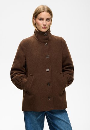 Femme aux cheveux clairs portant un manteau marron à col haut boutonné et un jean bleu, debout avec les mains dans les poches du manteau devant un fond uni.