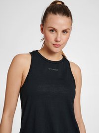 Junge Frau mit zurückgebundenen Haaren, die ein ärmelloses schwarzes Sporttop mit "hummel"-Logo auf der Brust trägt und in die Kamera schaut.