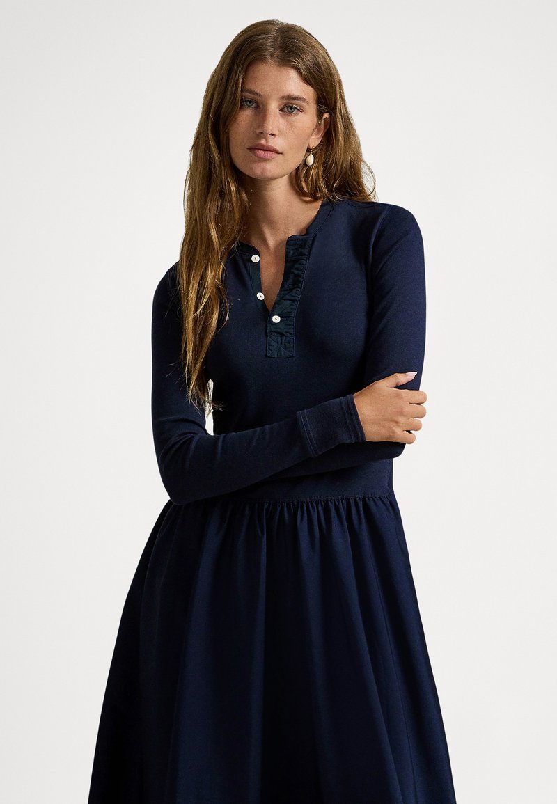 Μίντι φόρεμα σε ναυτικό μπλε με μακριά μανίκια, κουμπωτό γιακά με βολάν και φούστα με πιέτες. Κατασκευασμένο από απαλό ύφασμα, διαθέτει εφαρμοστό κορσάζ και ρέον κάτω μέρος.