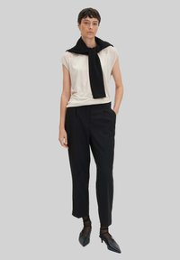 Beige Kurzarmoberteil, schwarzer gestrickter Schal um den Hals drapiert, schwarze maßgeschneiderte Hose und schwarze spitze Schuhe. Einfarbiger Hintergrund.