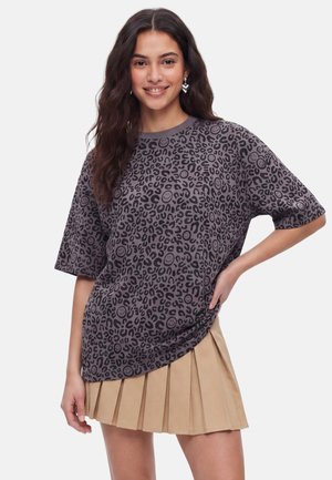 Ung kvinde med langt mørkt hår iført en grå leopardprint skjorte med smileys og et beige, plisseeet skørt, smilende med hånden på hoften.