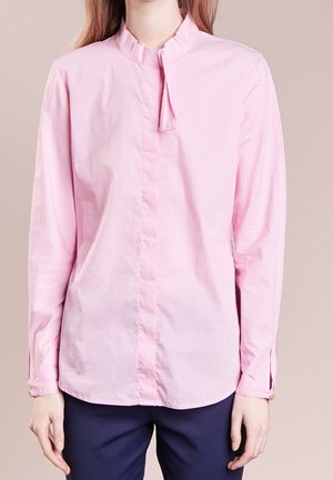 Frau trägt eine hellrosa Langarmbluse mit kleinem Nackenschleifen-Detail, kombiniert mit dunkelblauer Hose, vor einfarbigem Hintergrund.