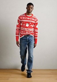 Roter Strickpullover mit weißen Schneeflocken und Weihnachtsmann-Gesichtern, kombiniert mit blauen Jeans und schwarzen Schuhen, auf einem Holzboden.