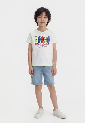 Boy wearing white t-shirt with colorful surfboards and "California Los Angeles" text, denim shorts, and white sneakers, standing still.