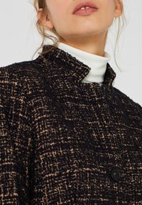 Chaqueta texturizada en negro y marrón con un alto cuello, que cuenta con un cierre de botón visible y un patrón de cuadros. Tejido suave y entrelazado.
