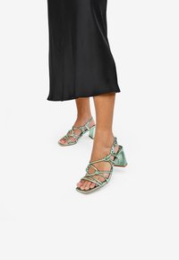 Sandalias de tacón verde con tiras, de punta cuadrada, que presentan un diseño abierto, detalles anudados y un tacón grueso. Acabado satinado.