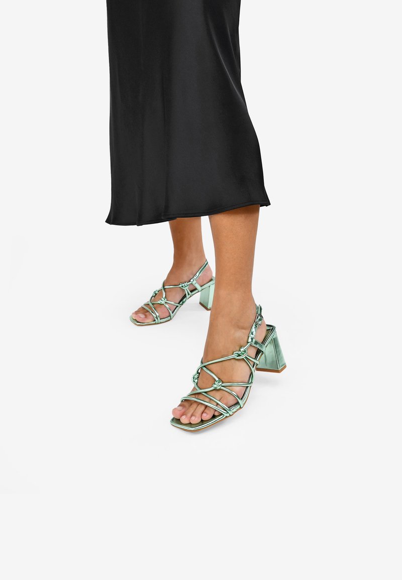 Sandalias de tacón verde con tiras, de punta cuadrada, que presentan un diseño abierto, detalles anudados y un tacón grueso. Acabado satinado.