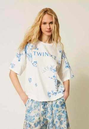 Giovane donna con lunghi capelli biondi che indossa una maglietta bianca oversize e pantaloni larghi, entrambi con motivi floreali blu.