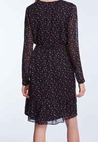 Zwart jurkje met lange mouwen, met een patroon van kleine rode en witte harten, een taille die is aangesnoerd en een gerimpelde zoom ter hoogte van de knie.