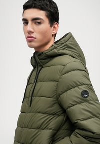 Zöld puffernadrág, steppelt mintázattal, csatolt kapucnival, elülső cipzárral és fekete húzózsinórokkal. Az ujján kör alakú logó található.