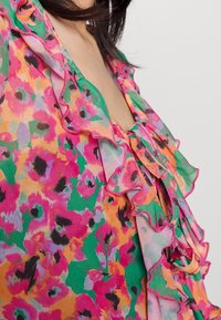 Blouse florale dans des teintes vives de rose, vert et orange. Présente un col à volants et un détail à nouer, fabriquée en tissu léger et texturé.