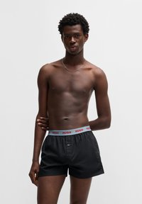 Sorte boxer shorts lavet af glat stof, med en grå elastik og rød "HUGO" tekst samt en knapåbning. Modellen fremstår i en neutral positur.