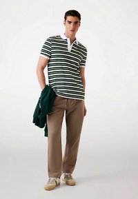 Polo majica kratkih rukava u zeleno-bijele pruge s bijelim ovratnikom. Bež hlače, svijetla obuća i zelena jakna prebačena preko ruke.