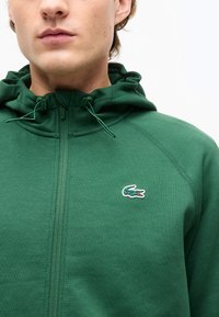 Άνδρας που φοράει πράσινο φούτερ με φερμουάρ, με ένα μικρό κεντημένο πράσινο λογότυπο κροκόδειλου στο στήθος κοντά στο φερμουάρ.