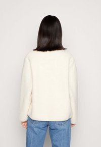 Kremowy sweter z polaru o prostym wzornictwie, z okrągłym dekoltem i długimi rękawami, zestawiony z jasnobrązowymi jeansami w kolorze jasnoniebieskim. Miękka tekstura i luźne dopasowanie.