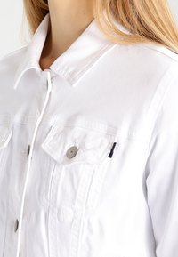 Valkoinen farkkutakki, jossa on terävä kauluskäänne, kaksi rintataskua, hopeiset napit ja teksturoitu kangas.