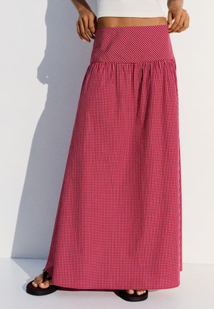 Femme portant une longue jupe rouge taille haute avec un petit motif à carreaux blancs et des sandales noires sur fond blanc.