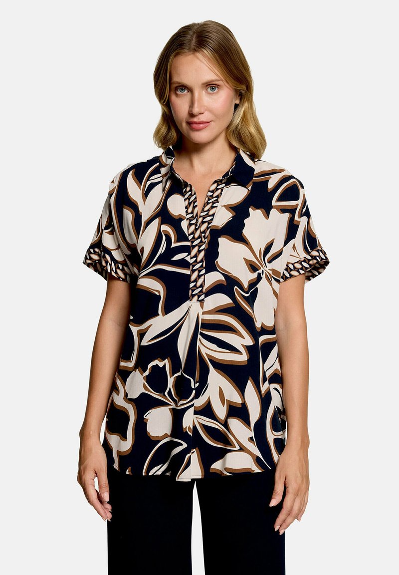 Femme portant un chemisier à manches courtes avec un grand imprimé floral noir, blanc et beige, debout les bras détendus le long du corps sur un fond uni.