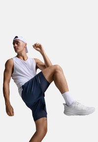 Biały sportowy top bez rękawów, granatowe szorty z panelami z siateczki i białe buty do biegania. Modelka pozuje z jedną nogą uniesioną, ubrana w czapkę.