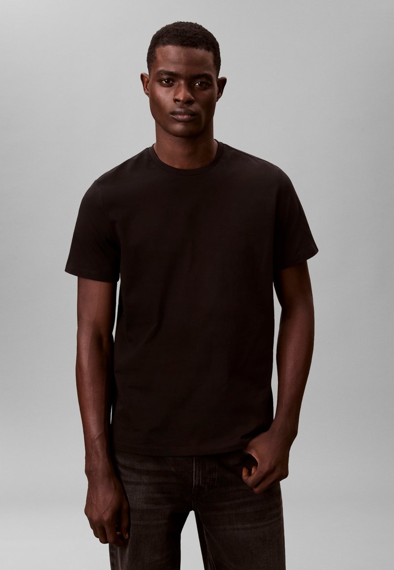 Zwart katoenen T-shirt met korte mouwen en een ronde hals, gecombineerd met donkere spijkerbroek, tegen een neutrale achtergrond. Strak, minimalistisch ontwerp.