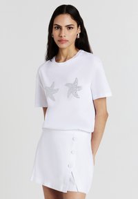 Femme aux longs cheveux noirs, portant une chemise à manches courtes blanche avec des motifs d'étoiles de mer argentées et une jupe blanche avec des boutons sur le côté, devant un fond uni.