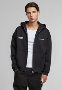 Felpa nera con cappuccio e zip, tasche frontali e testo logo bianco sul petto. Superficie liscia, vestibilità comoda e polsini in maglia.