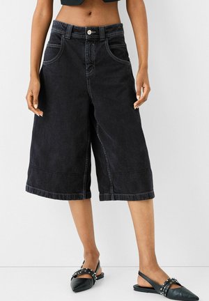 Culotte in denim nero con vestibilità ampia, cinque tasche, chiusura con bottone superiore e dettagli di cucitura in contrasto, abbinati a scarpe slingback nere.
