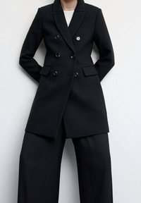 Persoon in een zwart dubbelbreasted blazer met zwarte wijde broek tegen een effen lichte achtergrond.