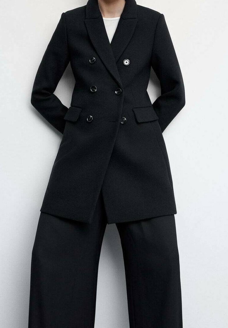 Persoon in een zwart dubbelbreasted blazer met zwarte wijde broek tegen een effen lichte achtergrond.