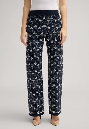 Pantalon large bleu marine avec motif géométrique et floral blanc, taille et poignets côtelés, porté avec des talons beige à bouts pointus et un haut blanc.