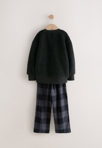 Sweat-shirt en polaire noir à surface texturée, associé à un pantalon ample à carreaux bleu marine et noir, suspendu à un cintre en bois.