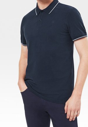 Granatowa koszulka polo z kołnierzykiem, białą lamówką i logo. Krótkie rękawy ukazują miękki, teksturowany materiał; noszona z ciemnymi spodniami, ręce w kieszeniach.