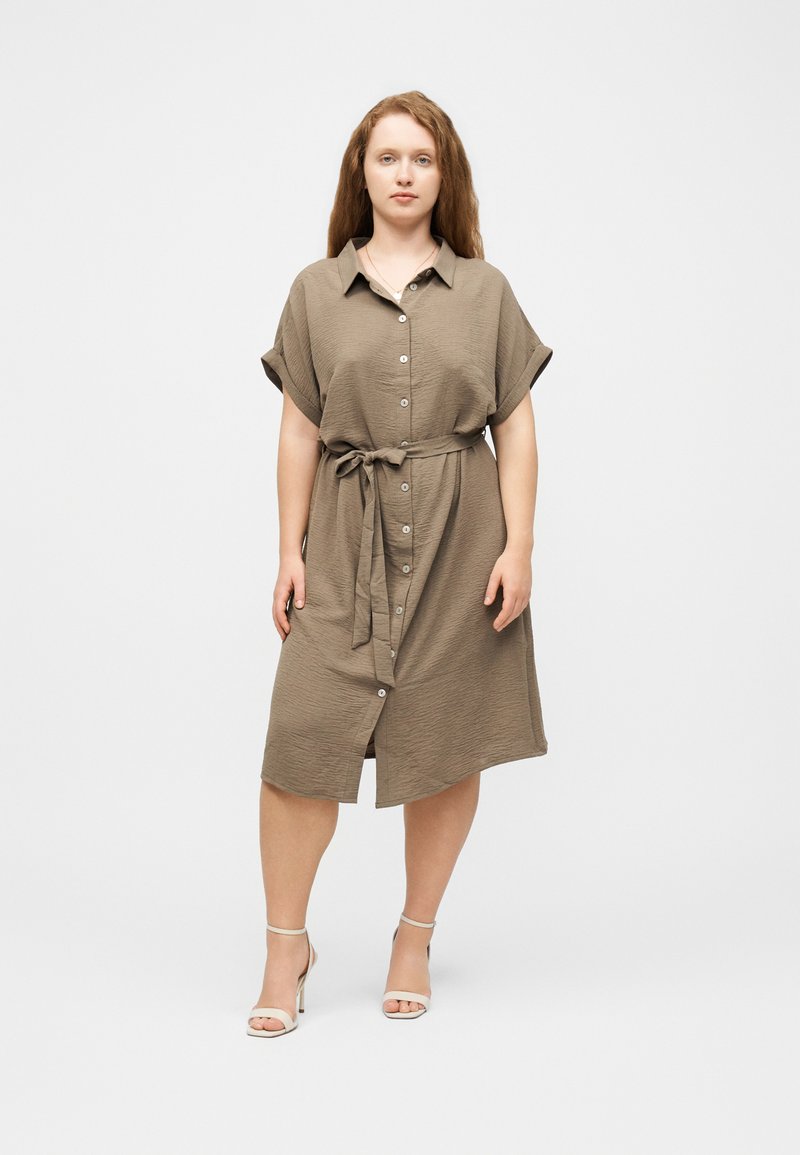 Γυναίκα στέκεται σε ένα καφέ φόρεμα μέχρι τα γόνατα με κουμπιά, κοντά μανίκια και δεμένη ζώνη στη μέση, φοράει λευκά πέδιλα με ψηλά τακούνια μπροστά σε απλό φόντο.