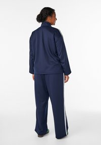 Námornícky športový komplet s vysokým golierom, dlhými rukávmi a bielymi bočnými pruhmi, obsahujúci voľné nohavice. Hladký, syntetický materiál s voľným strihom.