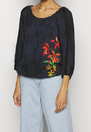 Kvinde iført en løs navyblå bluse med sorte blomsterprint og klare orange og gule blomster, kombineret med lyseblå jeans.