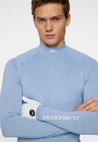 Vaaleansininen pitkähihaista paitaa, jossa on korkea kaulus ja joustavaa materiaalia. Kauluksessa on logo "JL" ja hihassa "J.Lindeberg".