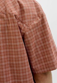 Camisa de manga corta a cuadros en color óxido con patrón de cuadrícula blanca. Presenta un corte relajado y una suave textura de algodón.