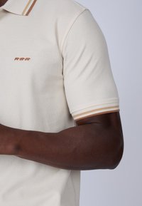 Polo crème en tissu texturé, avec une broderie de trois ours en rouille et des poignets rayés marron et beige.