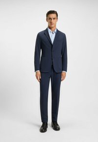 Completo blu navy con blazer a due bottoni, revers a punta e tasche applicate, abbinato a pantaloni coordinati. Il tessuto appare morbido con una minima lucentezza.