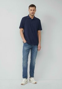 Granatowa koszulka polo z krótkim rękawem, w połączeniu z jasnoniebieskimi dżinsami i białymi trampkami z beżowymi akcentami i sznurowanym zapięciem.