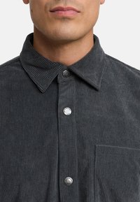 Camisa de pana gris con cuello extendido, botones plateados y un bolsillo en el pecho. La textura es acanalada, con líneas verticales sutiles.