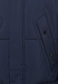 Merijääva puffatakki, jossa on tikattu muotoilu, vinossa etutaskussa on musta nappi ja sileä, kiiltävä kangaspinta.