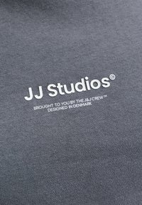 Tissu gris avec texte blanc imprimé "JJ Studios © Offert par l'équipe J&J™ Conçu au Danemark."